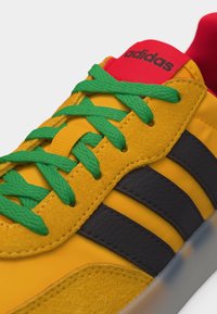 Κίτρινο αθλητικό παπούτσι Adidas με πράσινα κορδόνια, μαύρες ρίγες και κόκκινη γλώσσα που φέρει το λογότυπο Adidas, εμφανίζεται σε κοντινή λήψη.
