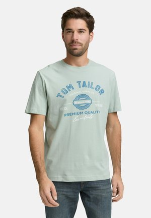 Mann trägt ein hellgrünes Tom Tailor Grafik-T-Shirt und blaue Jeans, steht vor einem schlichten weißen Hintergrund.