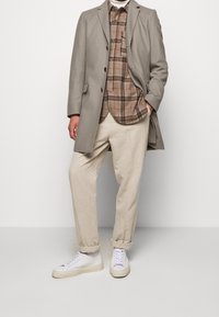 Cappotto grigio in lana, camicia a quadri marrone, pantaloni beige chiaro e sneakers bianche. Presenta bottoni e orli dei pantaloni arrotolati per un fit rilassato.