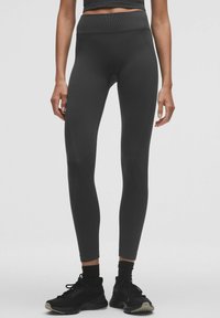 Hoge zwarte leggings van een gladde, elastische stof, met geometrische naden voor een aansluitend ontwerp. Gepaard met zwarte sneakers.