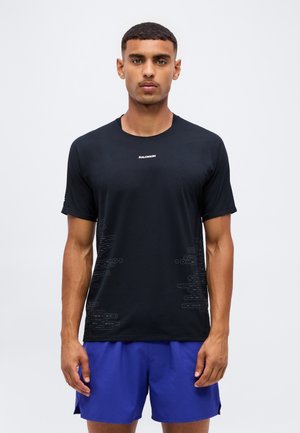 Joven con el pelo corto que lleva una camiseta negra de Salomon y pantalones cortos deportivos azules, de pie frente a un fondo liso y claro.