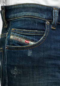 Jeans de mezclilla azul oscuro con una textura ligeramente desgastada, que presentan costuras naranjas y un parche de logo visible en el bolsillo delantero.