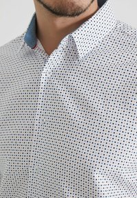 Homme portant une chemise blanche à boutons avec un petit motif de carrés bleus, noirs et bruns et un contraste de doublure de col bleu.