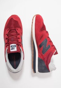 Pár červených tenisek New Balance 520 s černým logem, bílou mezipodešví, gumovou podešví a materiály z semiše a síťoviny na bílém pozadí.