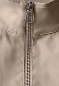 Tissu beige avec texture visible, comportant une fermeture éclair en métal argenté au centre, partiellement ouverte, et des bords cousus.