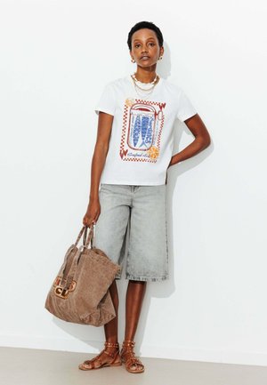 Femme portant un t-shirt graphique blanc, un short en jean gris jusqu'aux genoux, des sandales marron cloutées, tenant un grand sac cabas marron, debout contre un mur blanc.