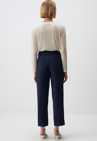 Leichte Creme lange Ärmel Top kombiniert mit hoch taillierten marineblauen Hosen und offenen weißen Schuhen, von hinten betrachtet.