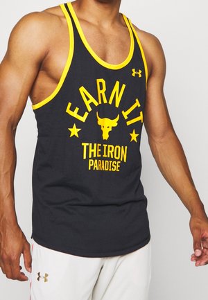 Muskelöser Mensch, der ein schwarzes Tanktop mit gelbem Rand und dem Text "Earn It The Iron Paradise" mit einem Stier-Logo trägt. Weiße Shorts mit Under Armour-Logo.