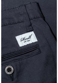 Primer plano de pantalones de tela oscura con una etiqueta blanca de la marca Reell y un botón metálico grabado con "REELL" cerca de un bolsillo trasero.