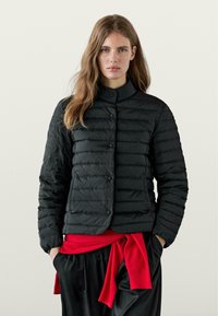 Giubbotto imbottito nero con colletto alto, caratterizzato da imbottitura orizzontale, bottoni a pressione e due tasche laterali. Una cintura rossa è legata in vita.