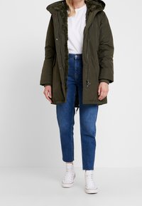 Olivově zelená parka s kapucí podloženou umělou kožešinou, zapínání na zip a boční kapsy, nošena přes světle modré džíny a bílé tenisky.