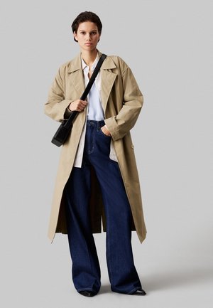 Vrouw draagt beige trenchcoat, wit overhemd, brede donkerblauwe spijkerbroek, zwarte puntige schoenen en zwarte schoudertas, staand tegen een grijze achtergrond.