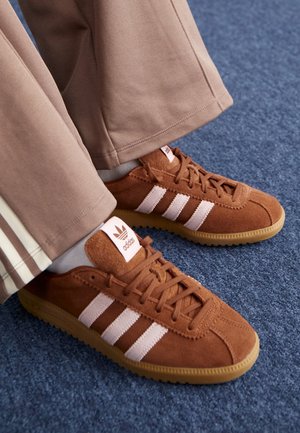 Hnědé semišové tenisky Adidas s bílými pruhy, nošené s světle hnědými kalhotami na modrém koberci.