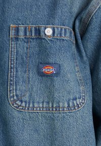 Denimskjorte med en brystlomme med orange syninger og en blå etiket, der viser "Dickies"-logoet i gule og røde farver.