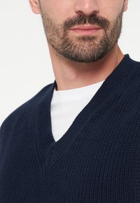 Homme barbu portant un pull en maille bleu foncé à col en V par-dessus un t-shirt blanc à col rond sur un fond uni.