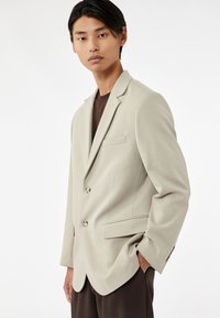 Lichtbeige blazer van geweven stof, met twee knopen, notch-kraag en een borstzak. Gecombineerd met een donkerbruine shirt eronder.