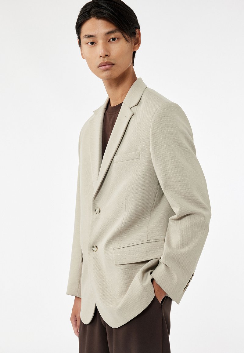 Lichtbeige blazer van geweven stof, met twee knopen, notch-kraag en een borstzak. Gecombineerd met een donkerbruine shirt eronder.