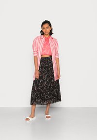 Top de renda cor-de-rosa cropped com uma camisa larga riscada, combinado com uma saia plissada preta que apresenta padrões de arte linear coloridos e sandálias brancas tipo slide.