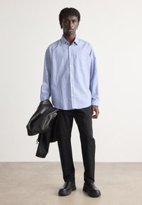Striped blue and white button-up shirt with a chest pocket, paired with black pants and a black leather jacket, standing on a neutral floor.