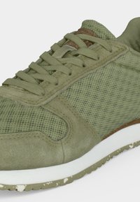 Zbliżenie na zielony meshowy i zamszowy sneaker z pasującymi sznurówkami oraz białą śródpodeszwą z teksturowaną gumową podeszwą zewnętrzną.