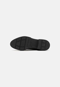 Semelle de chaussure noire avec un caoutchouc texturé, dotée de motifs de grip rectangulaires et du nom de la marque en relief. Fini lisse sur la partie du talon.