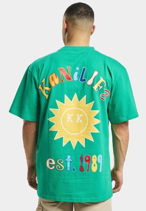 Green cotton t-shirt featuring colorful text and a yellow sun graphic on the back. Loose fit with short sleeves, casual design.