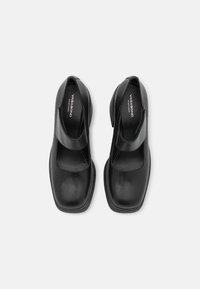 Chaussures style Mary Jane en cuir noir avec bouts carrés et brides à Velcro, portant la marque "Vagabond Shoemakers" à l'intérieur.