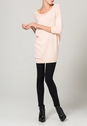 Světle růžový oversized svetr s korálkovým kulatým výstřihem, nošený přes černé skinny džíny a lesklé černé kotníkové boty.