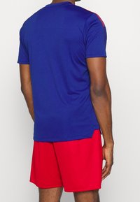 Modré sportovní tričko s červenými akcenty, krátkými rukávy a kulatým výstřihem, v kombinaci s červenými šortkami, představující hladkou, lehkou tkaninu.