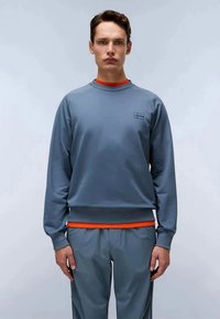 Jovem vestido com um sweat azul e calças combinando, com uma camisa laranja visível por baixo, encostado a um fundo claro e neutro.