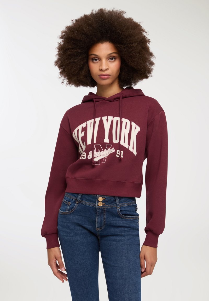 Bordowy krótki hoodie z białym napisem "NEW YORK" i logo. Posiada kaptur z regulacją i ściągacze przy mankietach. W połączeniu z niebieskimi dżinsami.