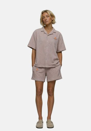 Femme aux cheveux blonds courts portant une chemise beige texturée à manches courtes assortie et un short, debout les mains dans les poches sur fond blanc.