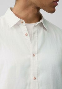 Chemise en lin blanche avec un col boutonné, deux boutons rose clair et une texture subtile. Portée par-dessus un sous-vêtement blanc.