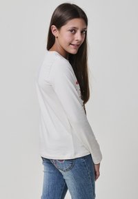 Junges Mädchen mit langen braunen Haaren, das ein weißes Langarmshirt und eine blaue Jeans trägt und mit einem leichten Lächeln über die Schulter zurückblickt.