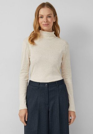 Kobieta z długimi blond włosami, ubrana w jasnobeżowy sweter z golfem oraz ciemnoszare, wąskie spodnie w prążki, stojąca na jednolitym tle.