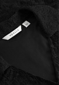 Čierna čipkovaná látka hladkého čierneho podšívky a dve biele oblečené štítky, z ktorých jeden zobrazuje "NOELLA" a druhý "S vyrobené v Číne."