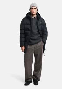 Sort pufferjakke, tekstureret grå sweater, ternet skjorte, brun plaid bukser og grå strikbeanie. Sorte sko fuldender outfittet.