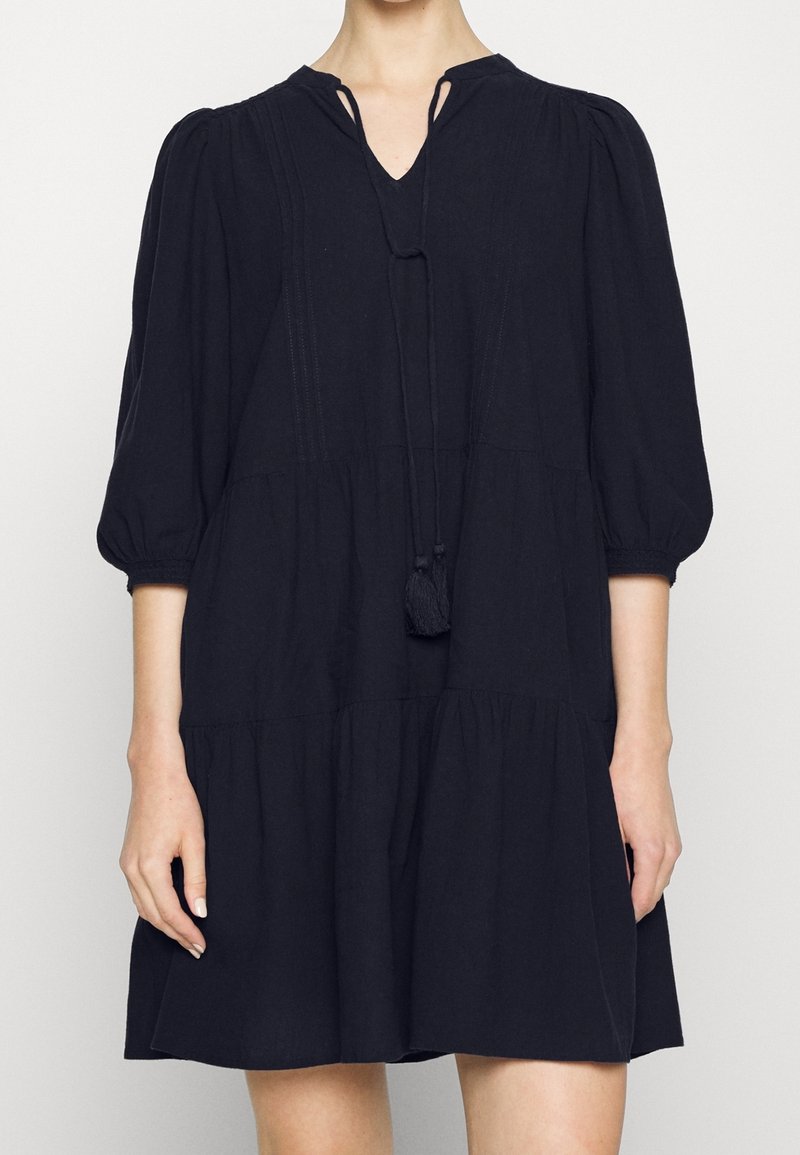 Femme portant une robe noire ample, longueur genoux, avec des manches ballon trois-quarts et un lien à l'avant avec des pompons au niveau de l'encolure.