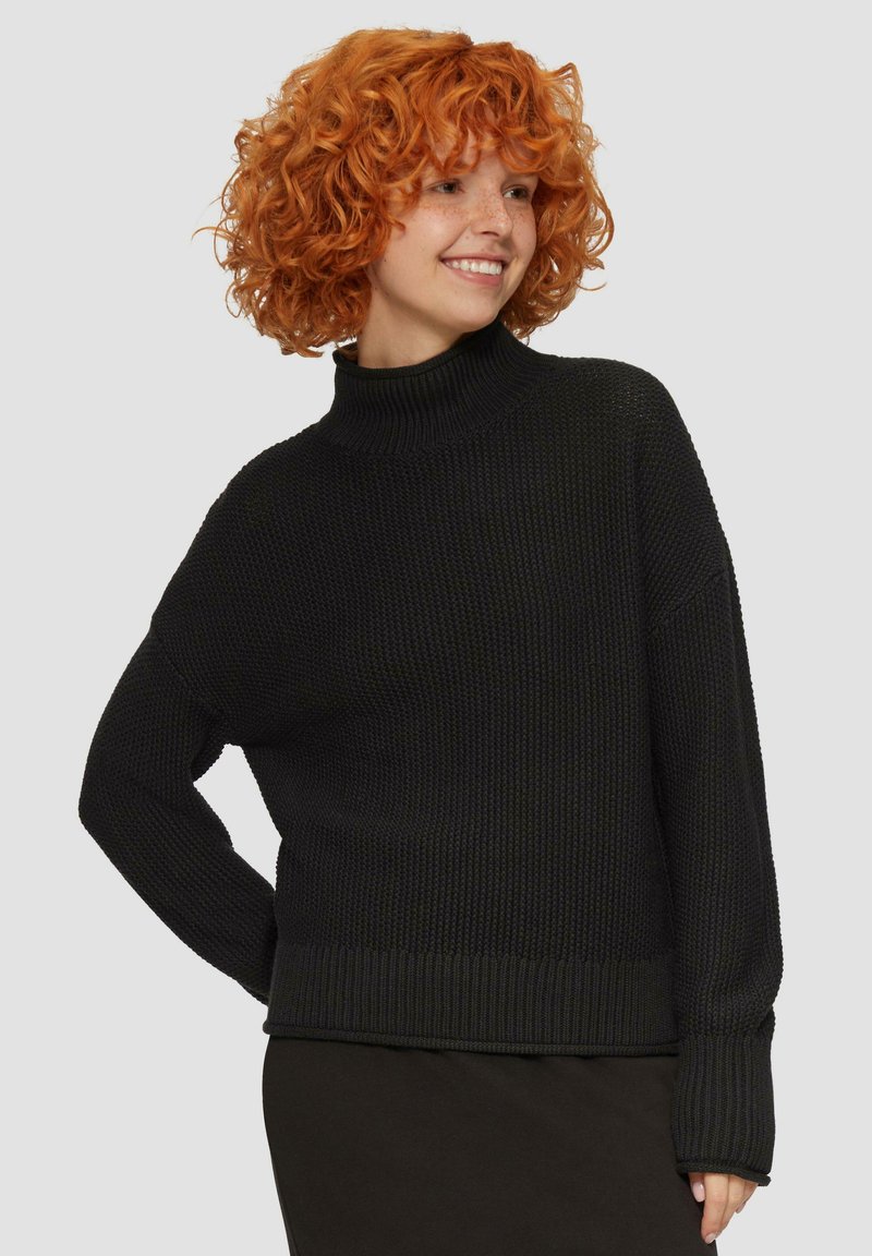Pull en tricot noir avec un col roulé haut, des épaules tombantes et des poignets côtelés. Surface texturée avec un design ample.