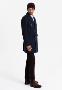 Cappotto doppiopetto blu navy in misto lana con bottoni dorati, abbinato a pantaloni slim-fit marroni e stivaletti alla caviglia marroni.