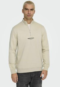 Sweatshirt beige zippé avec un col montant, arborant le texte imprimé "CURATED STAPLES" sur la poitrine, fabriqué dans un matériau doux. Jean gris.