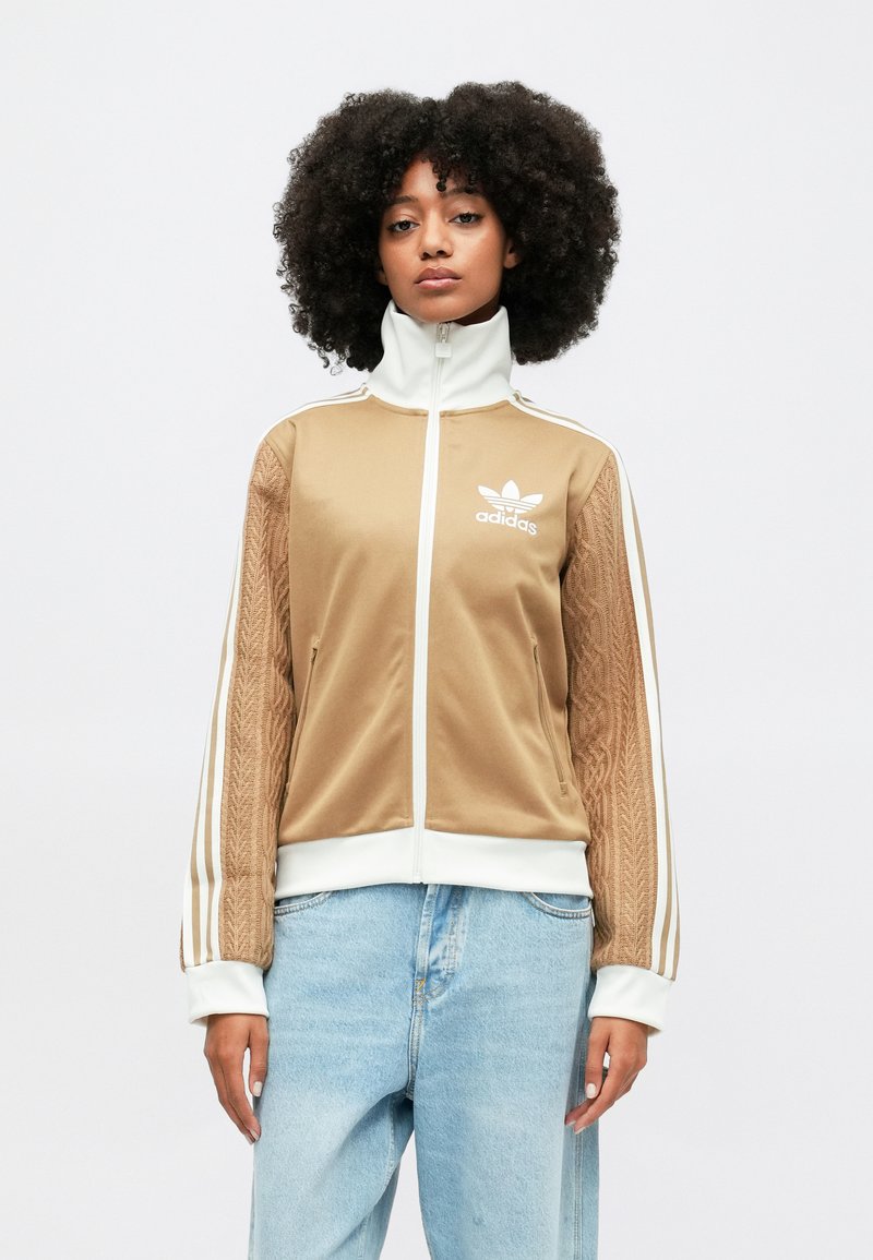 Braune, zip-up Sportjacke mit strukturierten Ärmeln, weißen Streifen, besticktem Logo, Stehkragen und Seitentaschen. Getragen mit hellen Jeans.