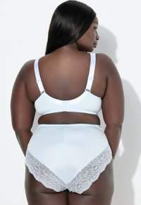 En kurvig kvinna med mörk hud bär ljusblå rippad lingerie med spetsdetaljer, sedd bakifrån mot en vit bakgrund.