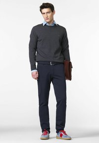 Grå sweater over en lyseblå skjorte, navybukser, rød og lyseblå sneakers, holder en brun lædertaske, med en ensfarvet hvid baggrund.