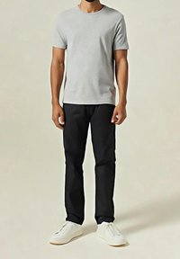 Homme portant un t-shirt gris clair à manches courtes, pantalon noir et baskets blanches, debout devant un fond clair uni.