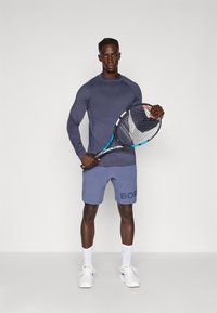 Langærmet navy atletisk skjorte, lyse shorts med "BOR" tekst, holder en tennisracket, hvide sneakers, ensfarvet baggrund.