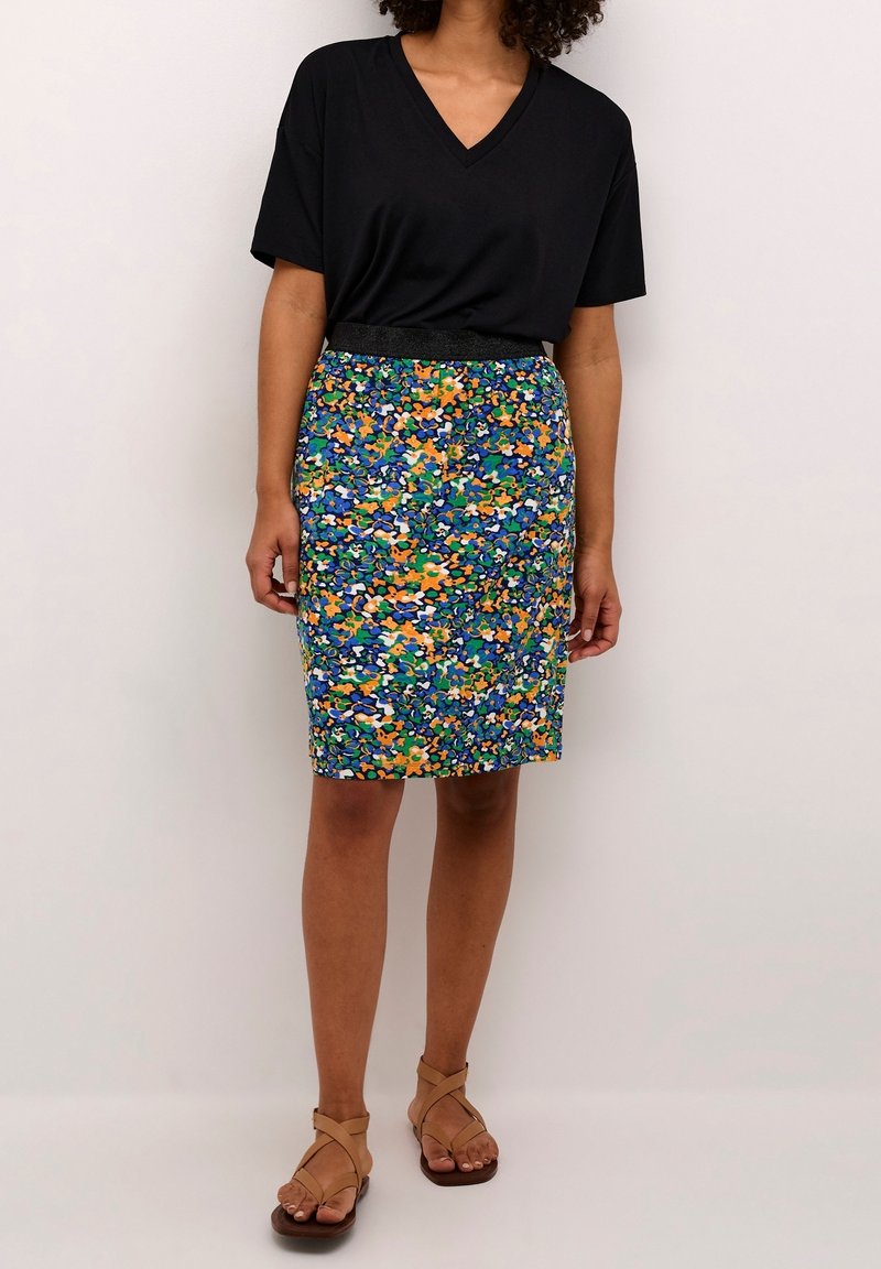 Femme portant un T-shirt noir à col en V, une jupe multicolore à fleurs arrivant aux genoux et des sandales plates beiges à lanières, debout devant un fond uni.