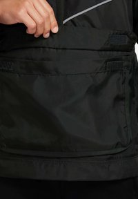 Czarna kurtka przeciwwiatrowa z dużą kieszenią z przodu z zapięciem na rzepy, gładką teksturą oraz odblaskowymi akcentami w okolicy zamka.
