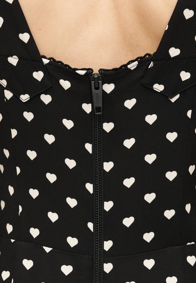 Κοντινό πλάνο μαύρου ενδύματος με λευκά μοτίβα καρδιάς και κεντρικό μαύρο φερμουάρ, με γιακά και κυματιστή άκρη κοντά στη λαιμόκοψη.