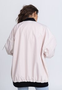 Veste bomber rose clair avec un ourlet noir en côtes, des poignets élastiques et une texture lisse et brillante. Présente une coupe décontractée et des fentes latérales.